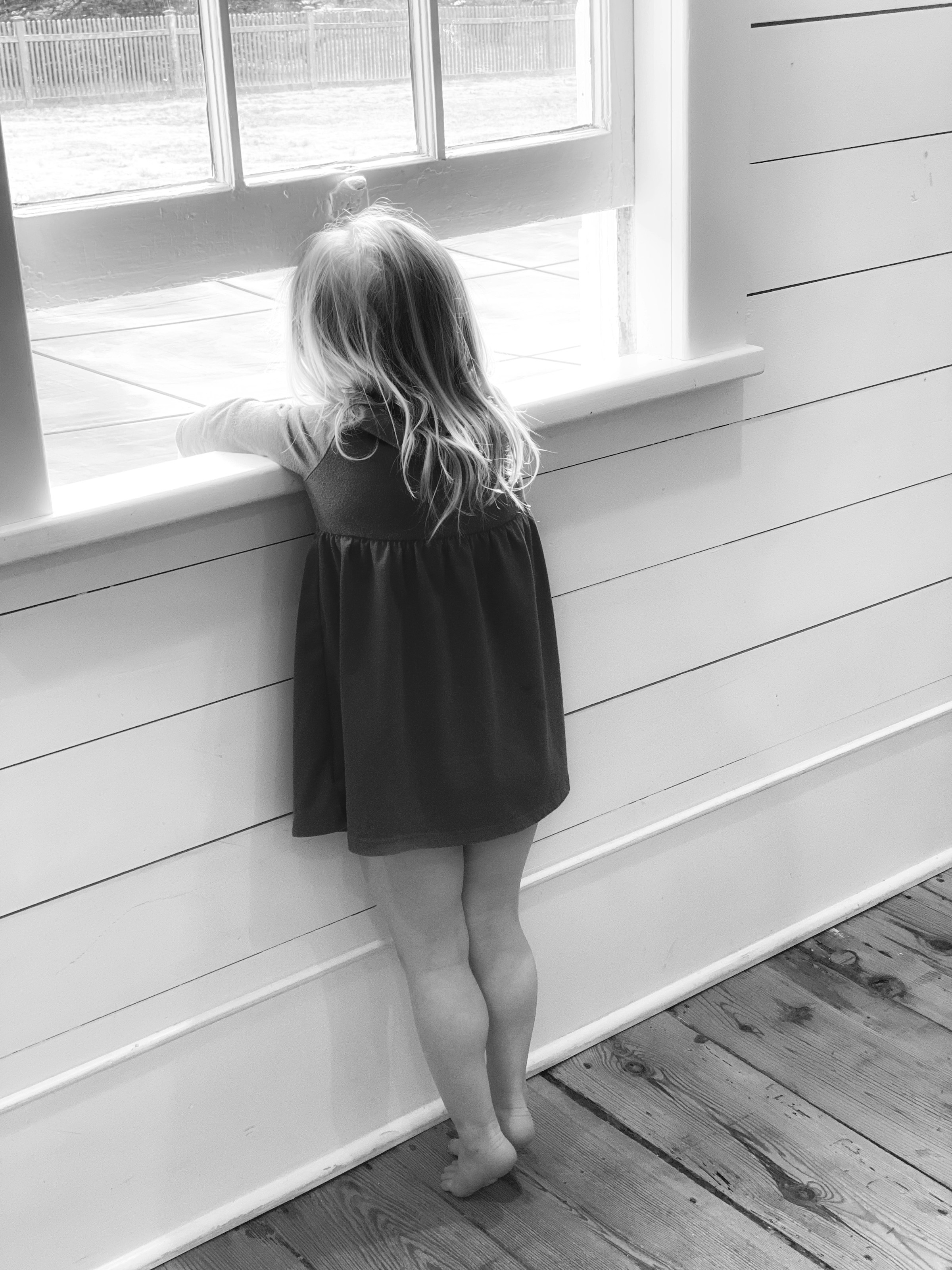 A grayscale shot of a little girl standing barefoot and looking out the window | Source: Unsplash