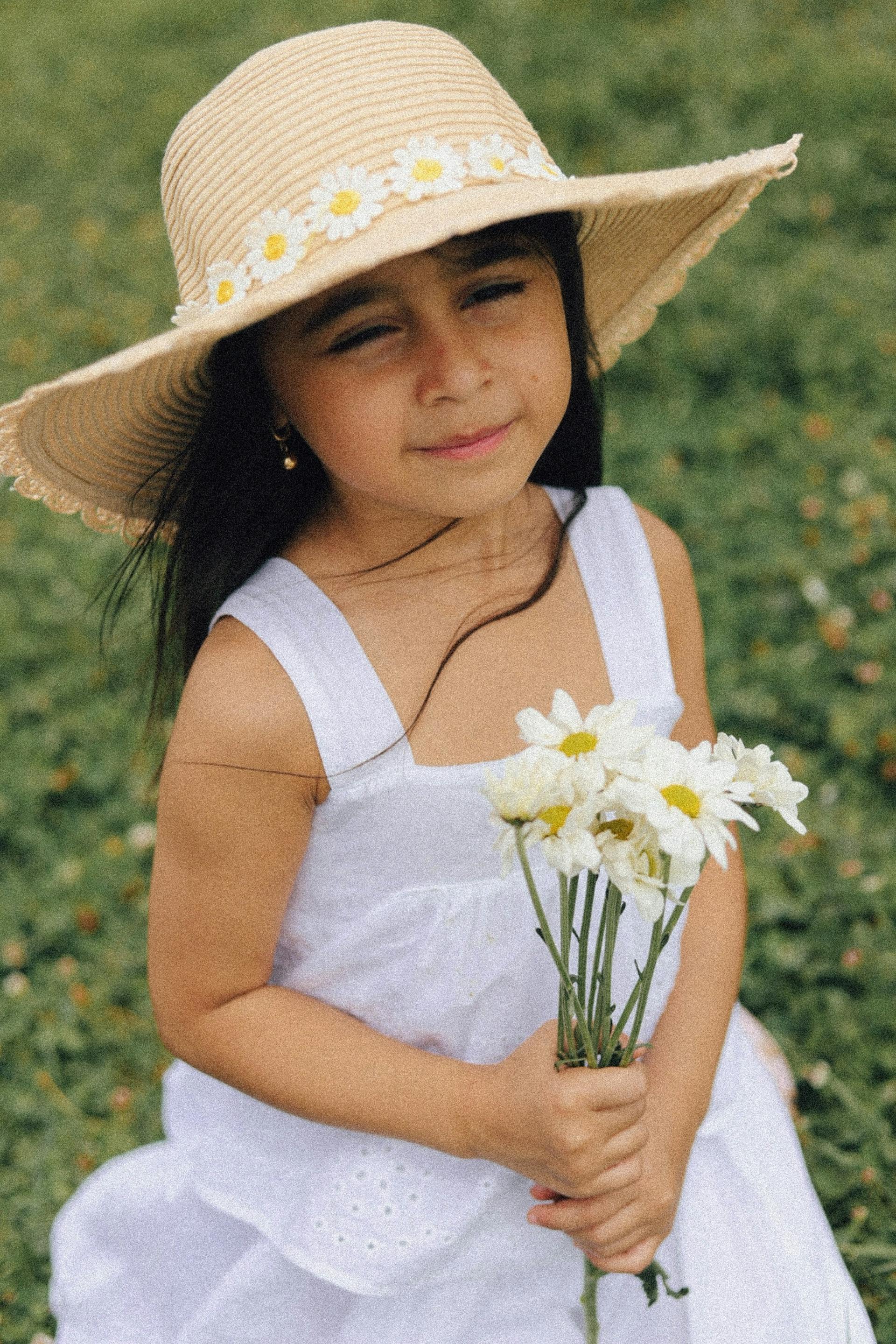 A little girl in a white dress and a straw hat with daisies holding a bouquet of flowers | Source: Pexels
