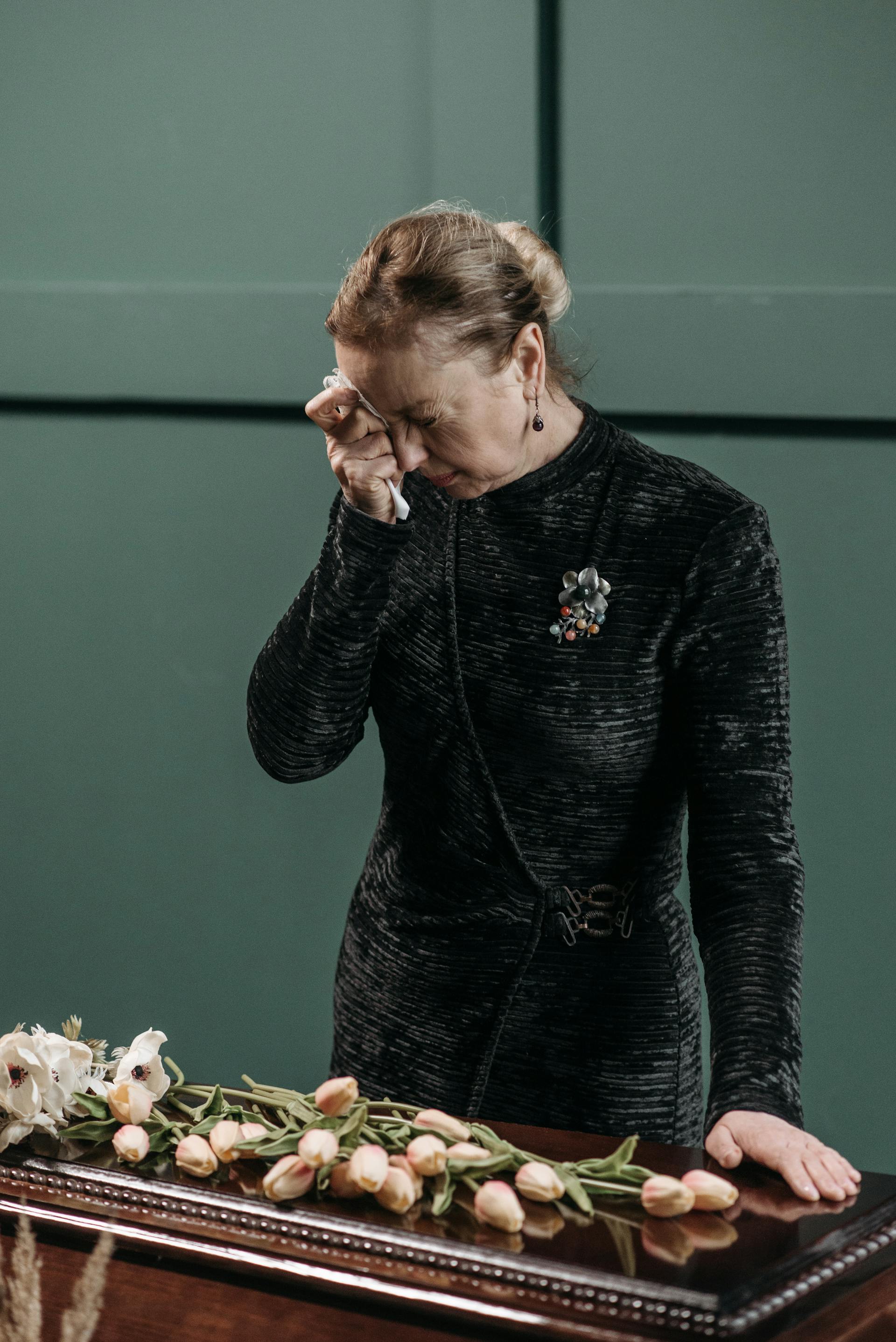A crying mature woman in a black dress wiping a tear with a tissue while standing by a casket | Source: Pexels