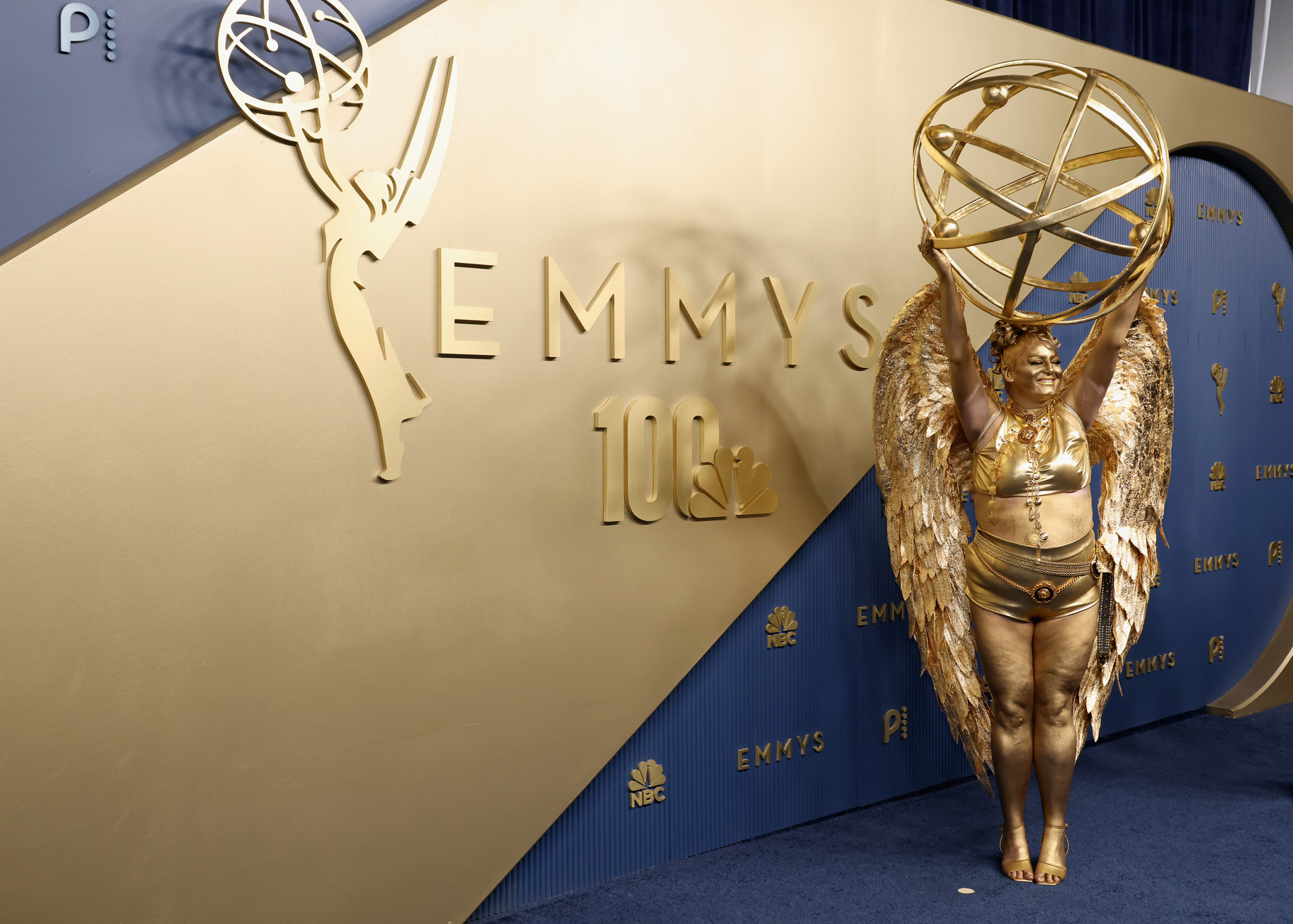 Meg Stalter poses in her metallic gold ensemble and sculpted wings against the Emmy Awards’ gold-and-blue backdrop, showcasing her elaborate hairstyle and golden body makeup. | Source: Getty Images