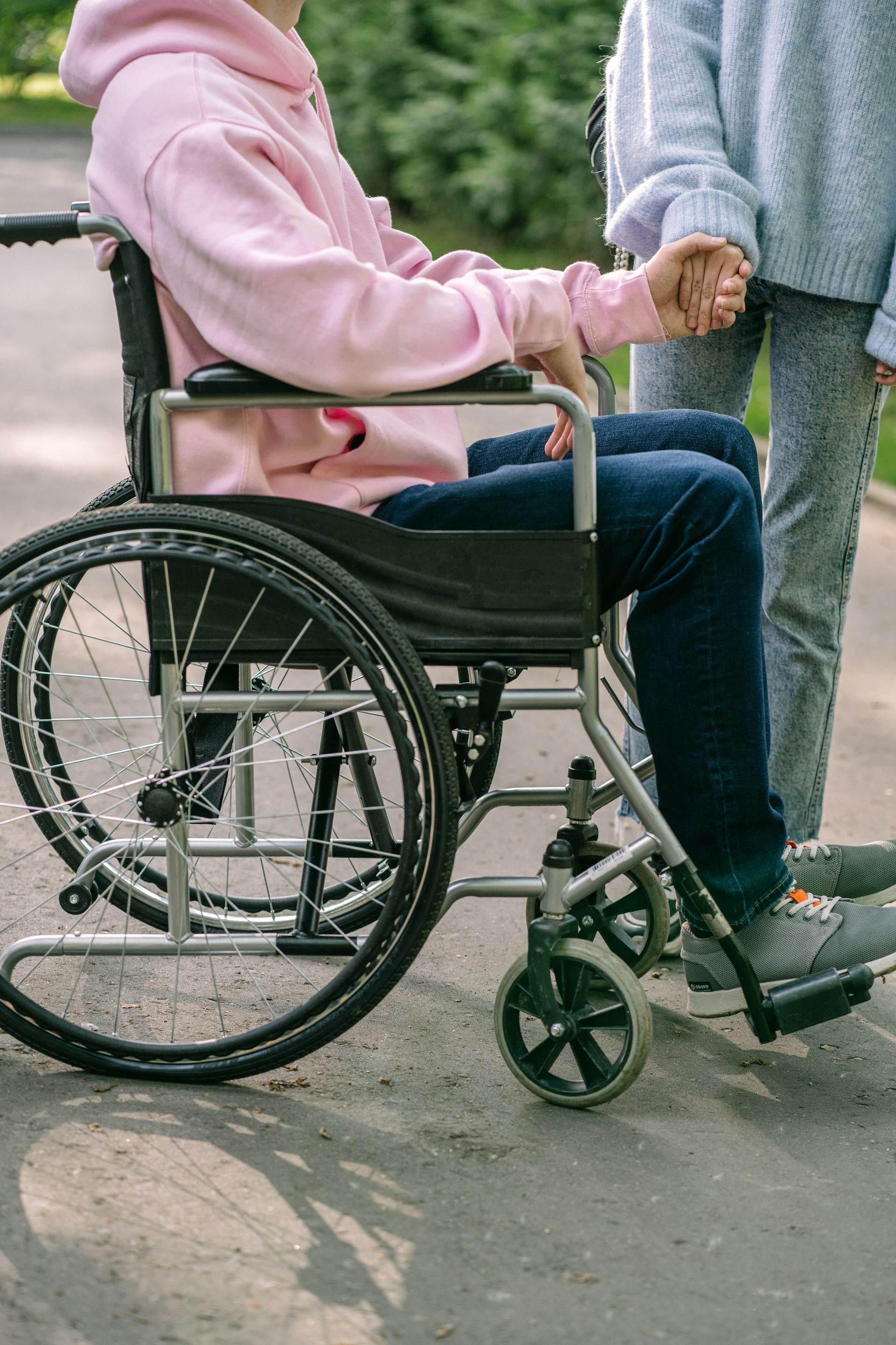A man in a pink hoodie sitting in a wheelchair and holding a woman's hand | Source: Pexels