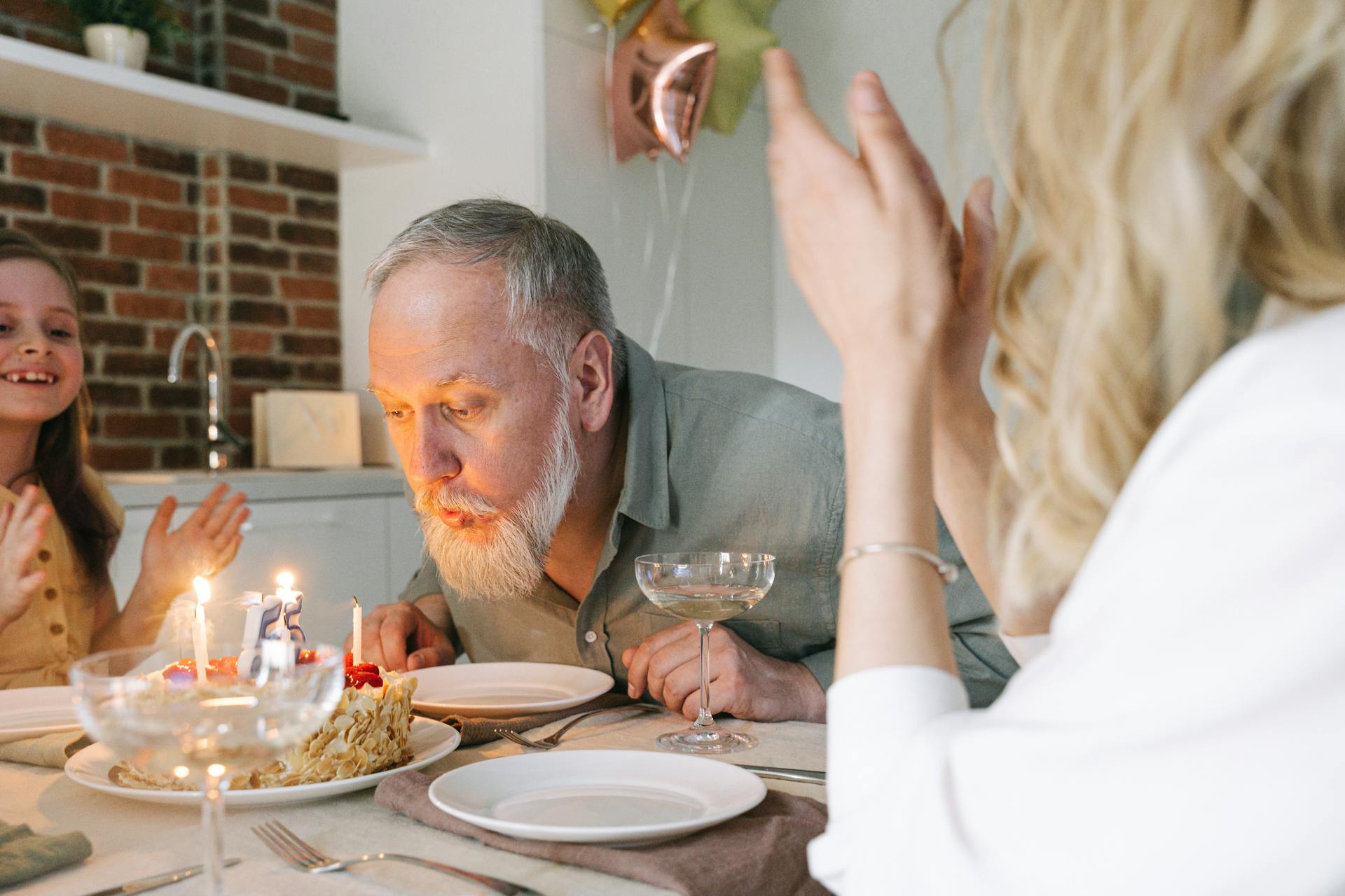 A mature man with a white beard blowing out candles on a birthday cake with family cheering him on | Source: Pexels