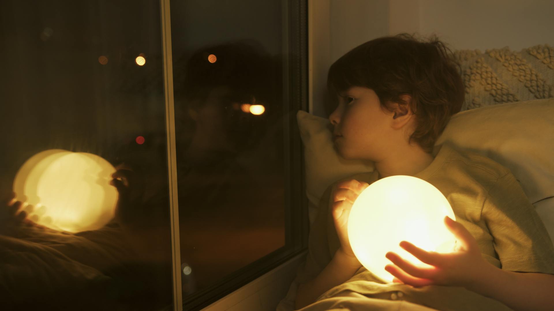 A young boy holding a ball of light peeking through his window at night | Source: Pexels