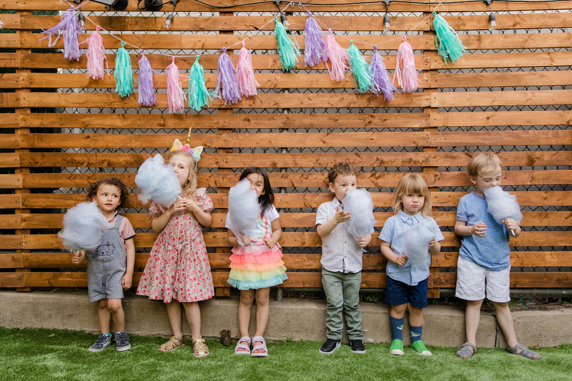 Kids eating cotton candies while standing in front of a wooden backdrop decorated for a party | Source: Pexels