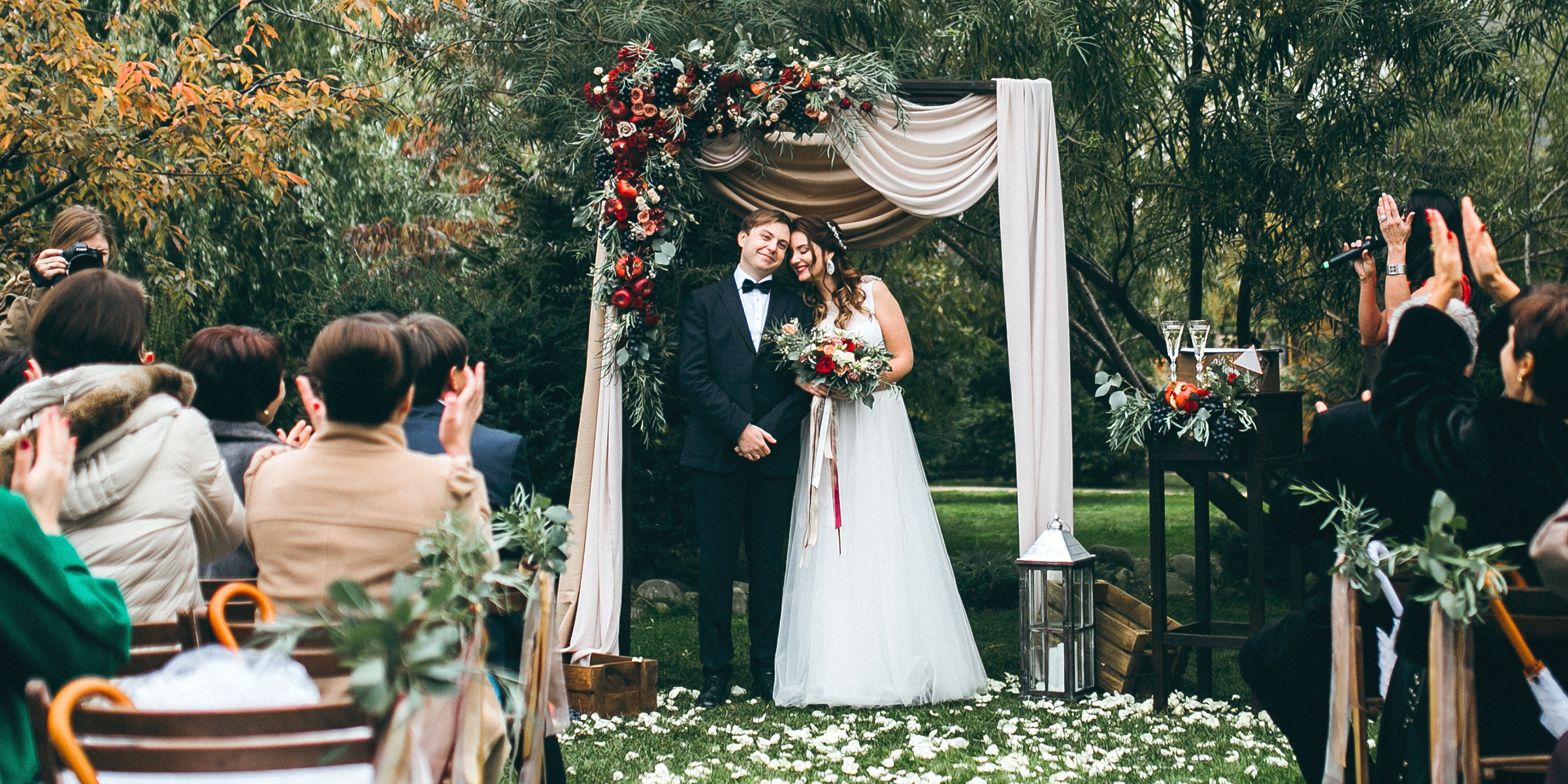 A bride and groom standing under a floral arch and smiling as guests cheer during their outdoor wedding ceremony | Source: Shutterstock