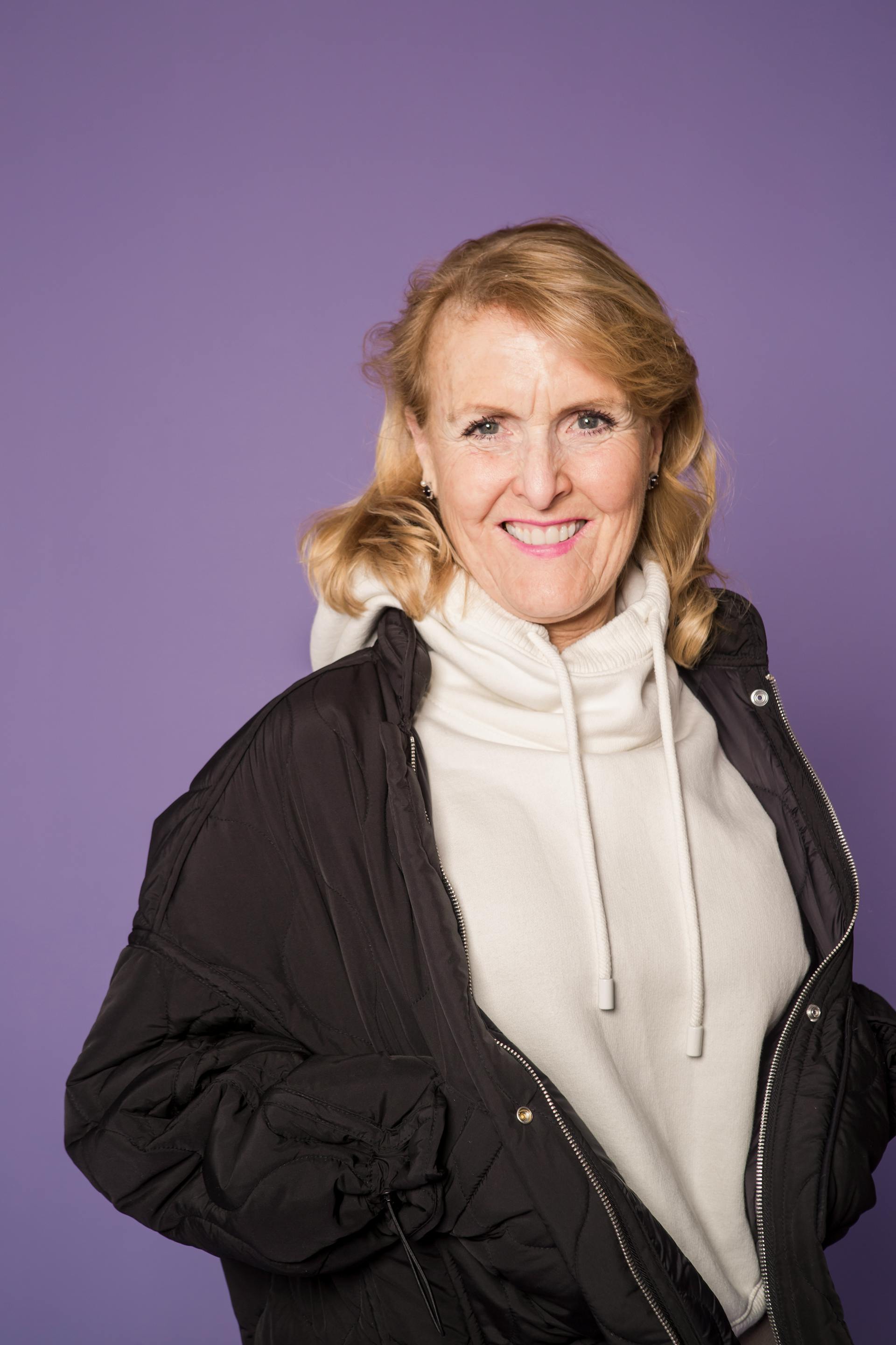 A happy mature woman in a black jacket and a white hoodie smiling against a purple background | Source: Pexels