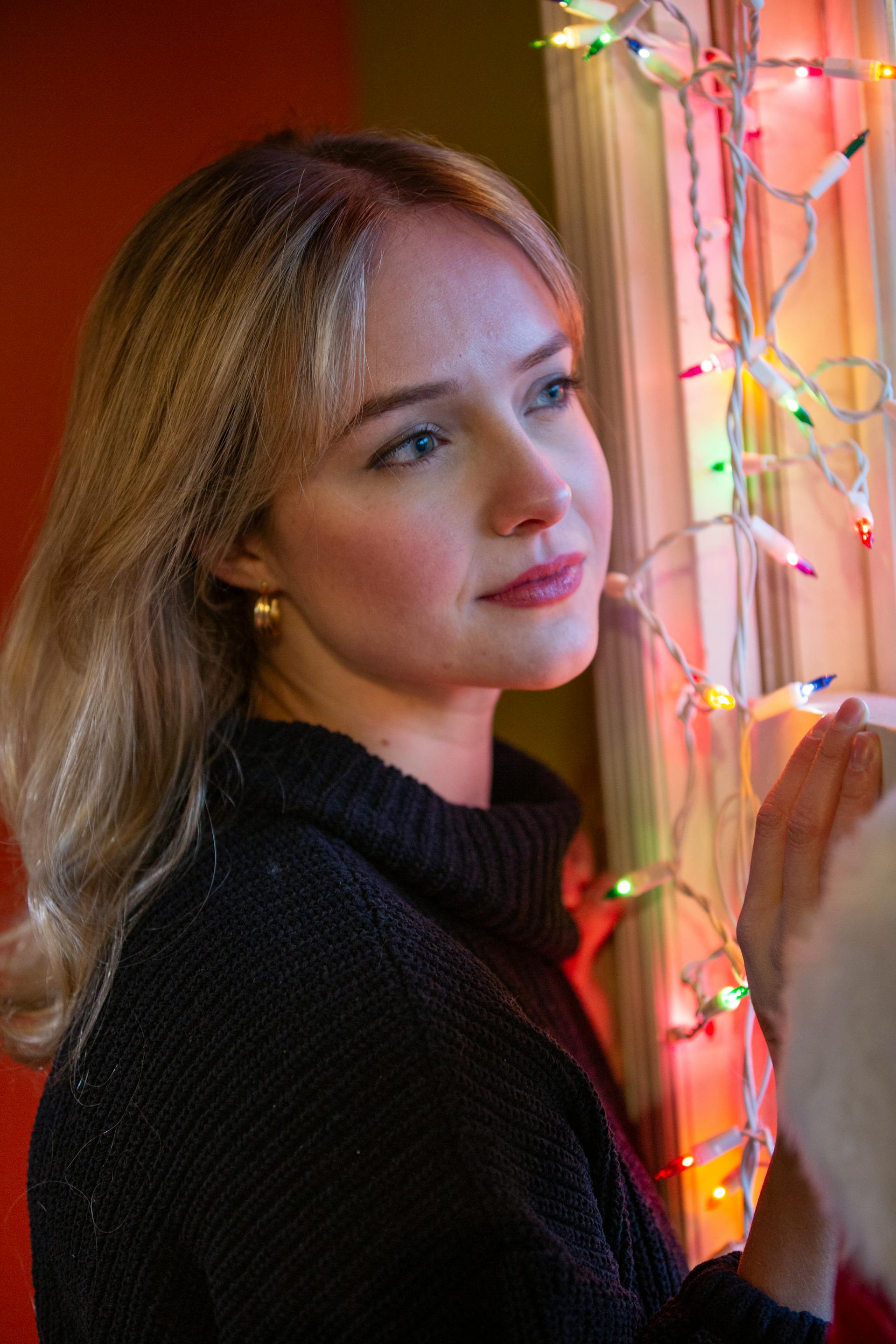 Contemplative woman standing by a window decorated with festive lights | Source: Pexels