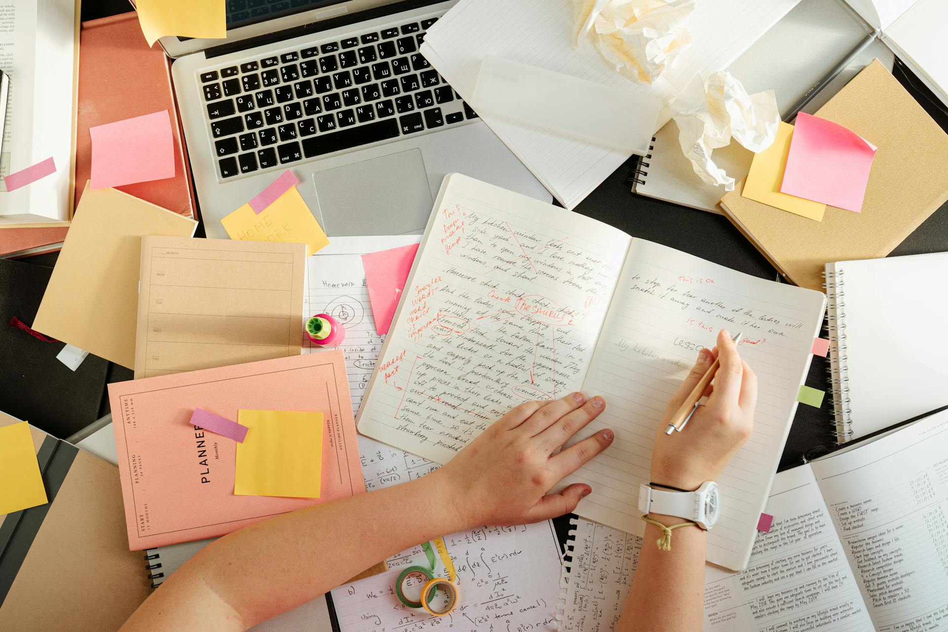 A messy desk with books, a laptop, and a person writing in a notebook | Source: Pexels