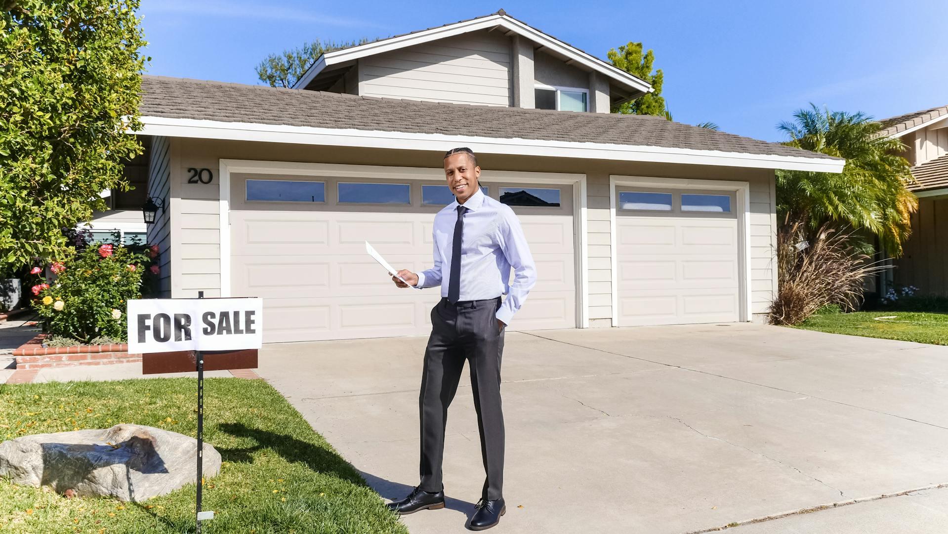 A smiling man standing in front of a house with a for sale sign | Source: Pexels