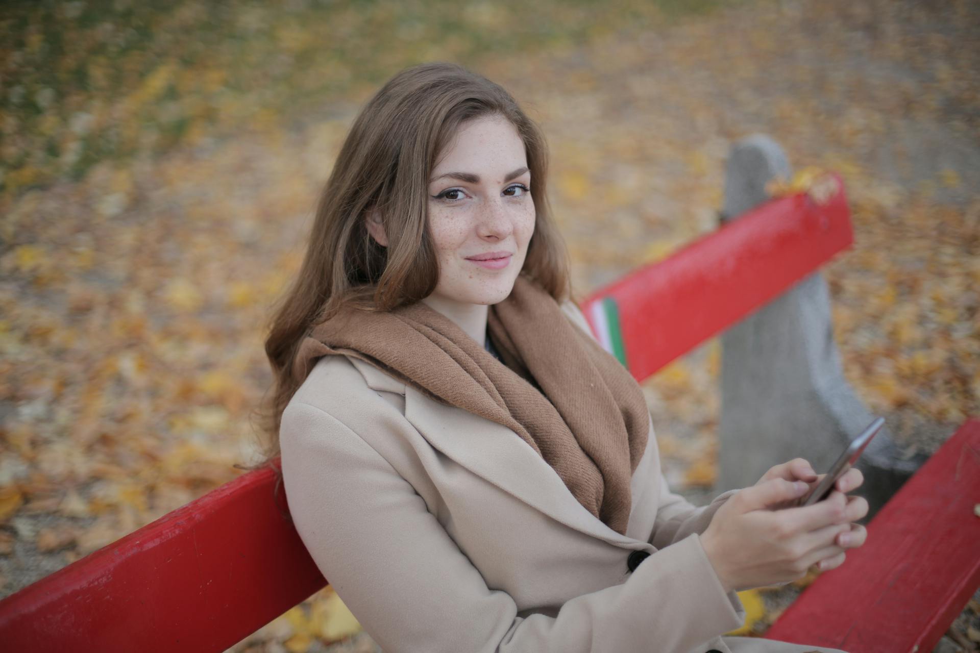 A woman in a coat sitting on a bench and holding her phone | Source: Pexels