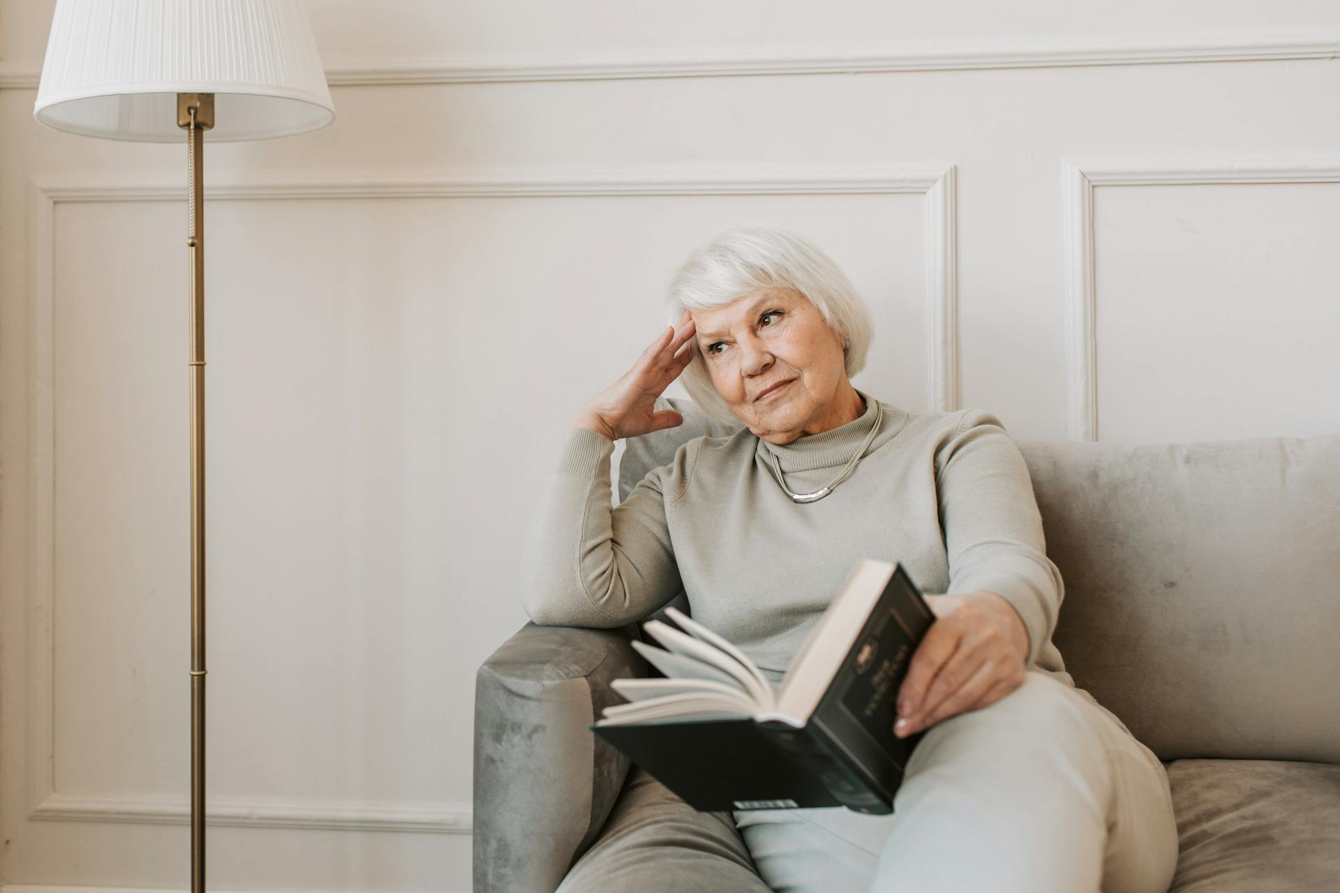 A pensive elderly woman sitting on a couch with a book | Source: Pexels