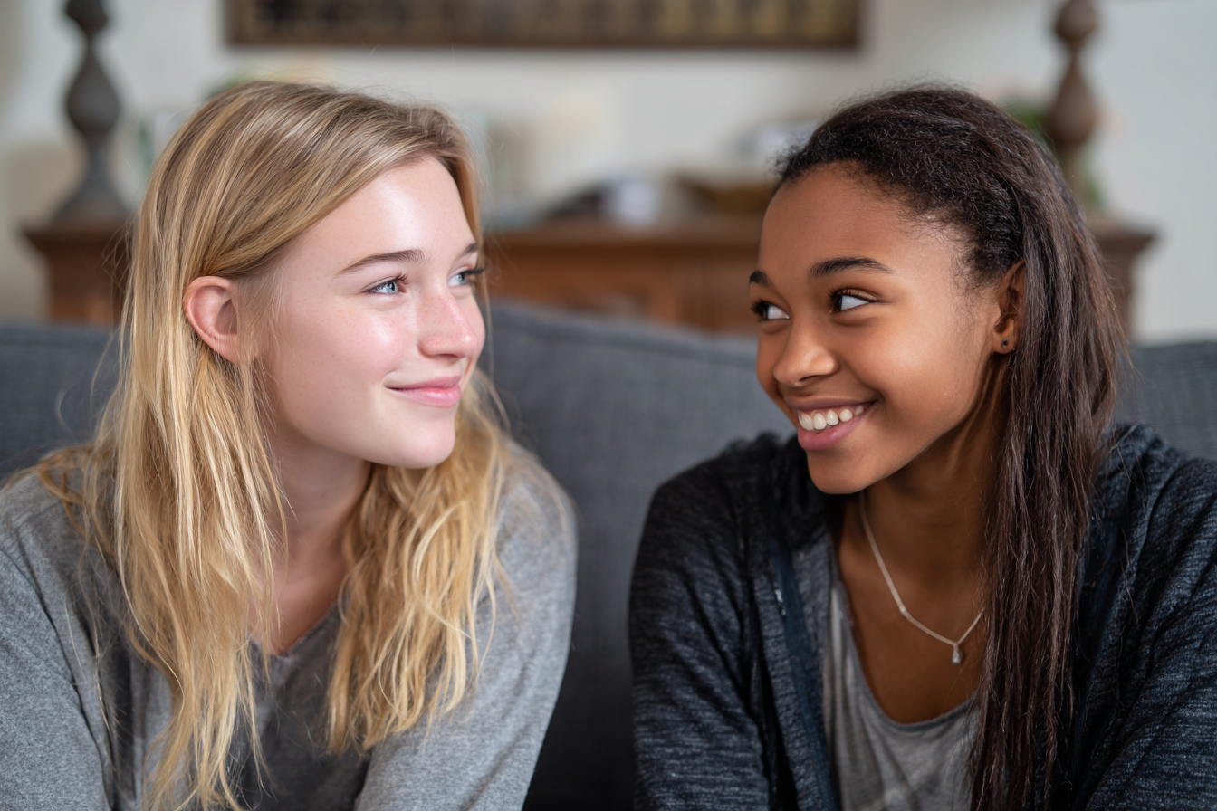 Two teen girls smiling at each other on a sofa | Source: Midjourney