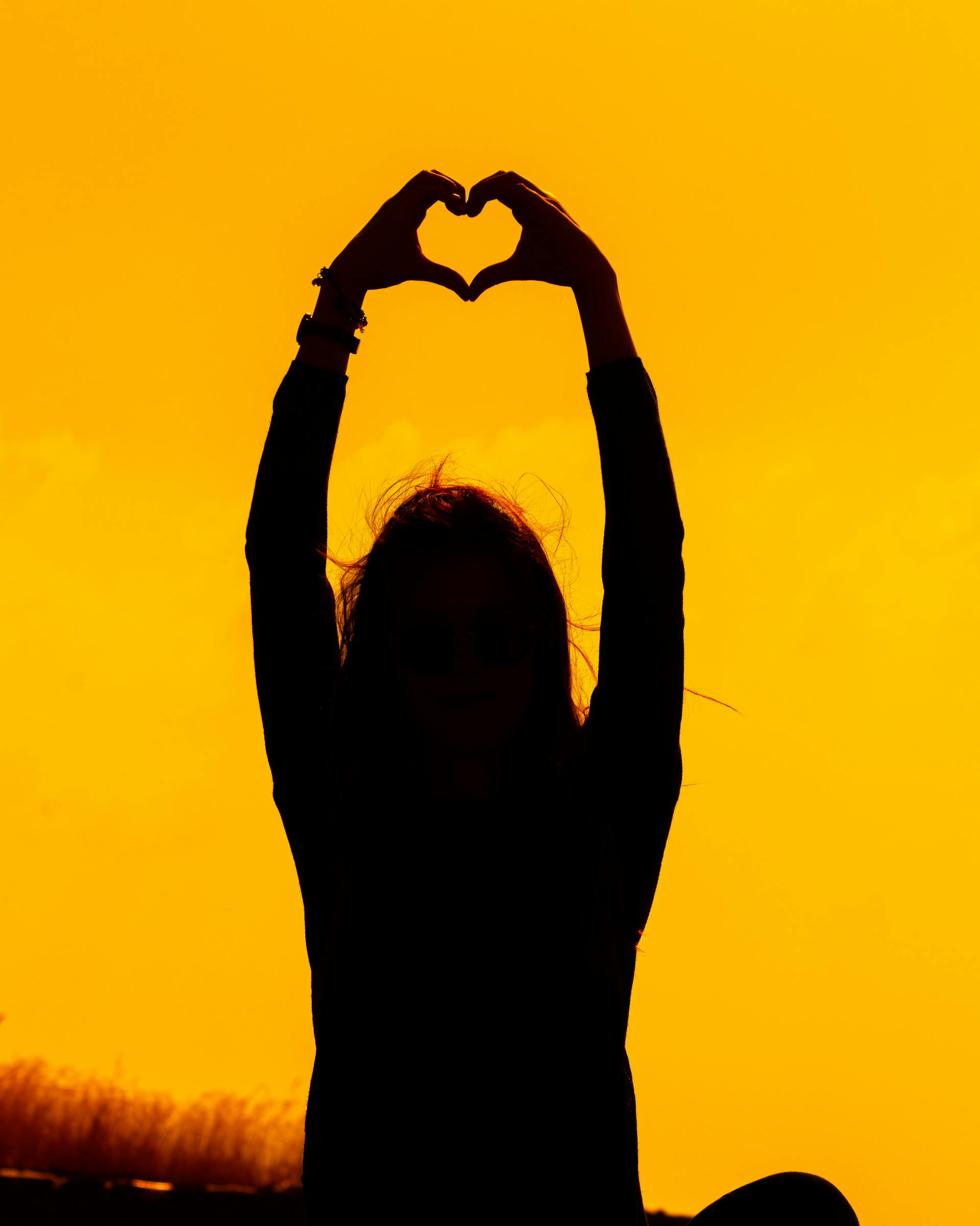 Sylwetka kobiety układająca dłońmi kształt serca o zachodzie słońca | Źródło: Pexels