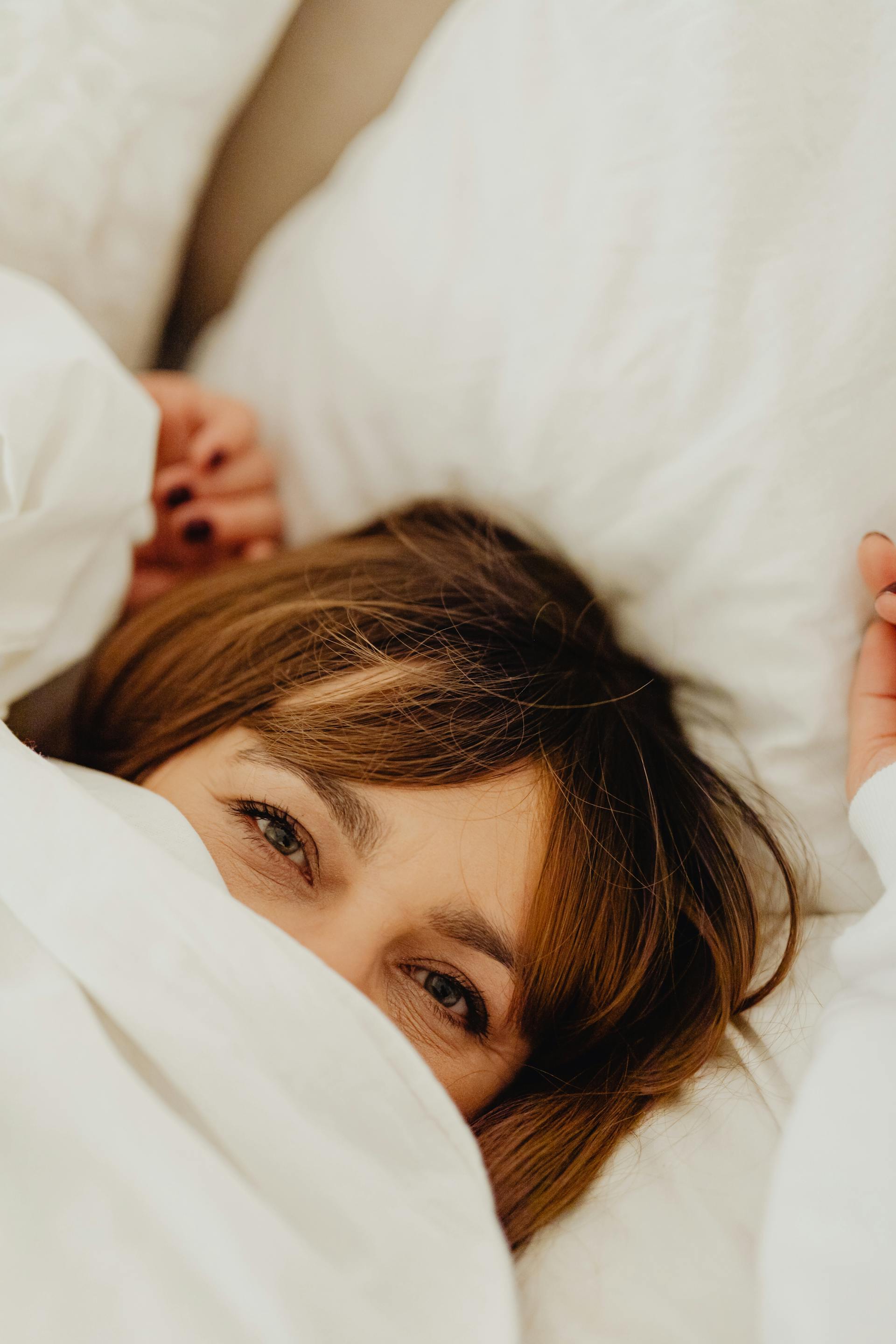 A woman lying in bed with her face hidden under a white blanket | Source: Pexels