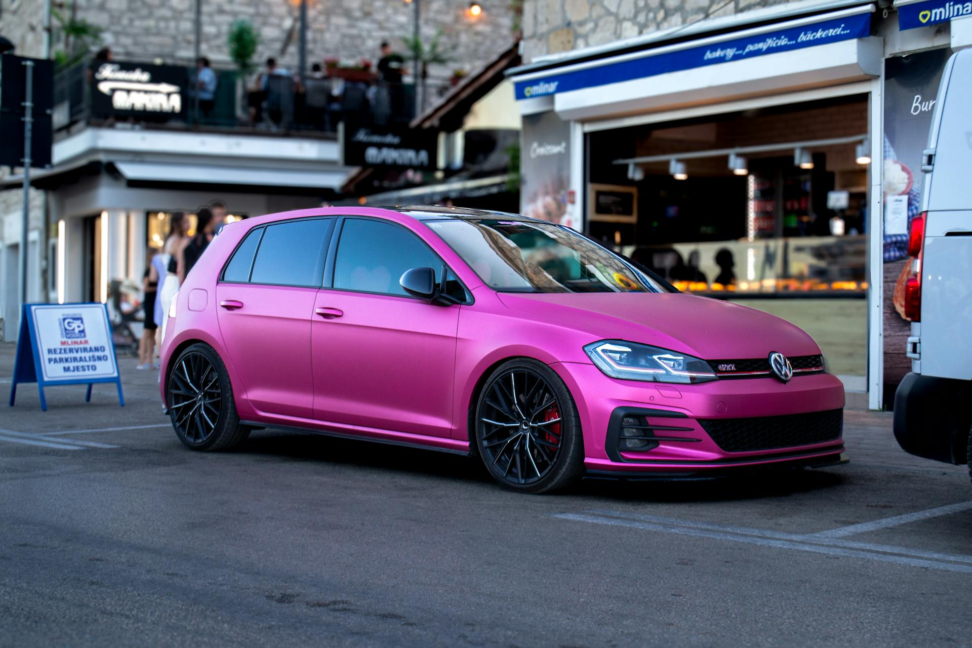 A matte pink hatchback car parked on a street with buildings and people in the background | Source: Pexels