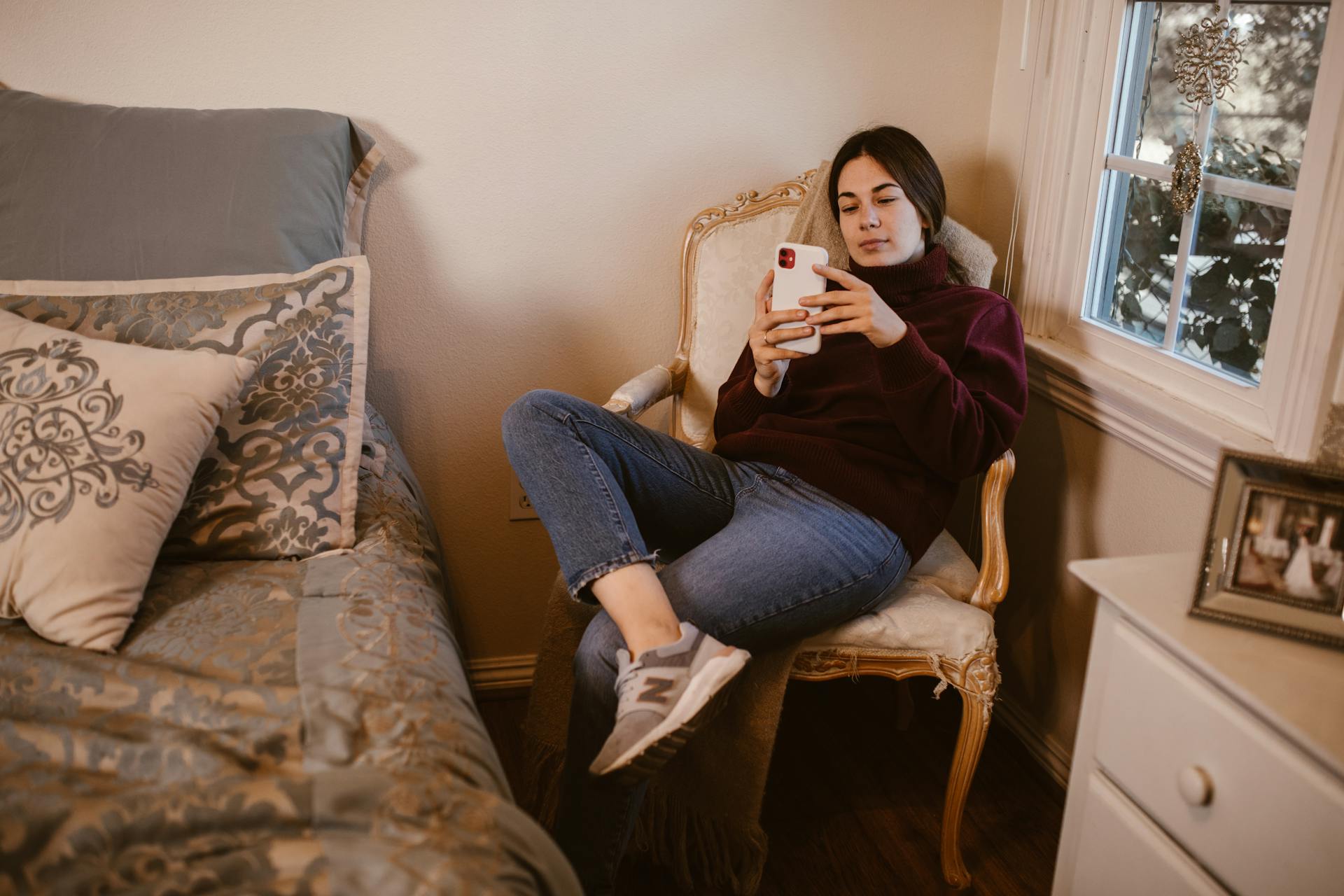 A young woman in a maroon sweater and jeans sitting in an armchair and looking at her phone | Source: Pexels
