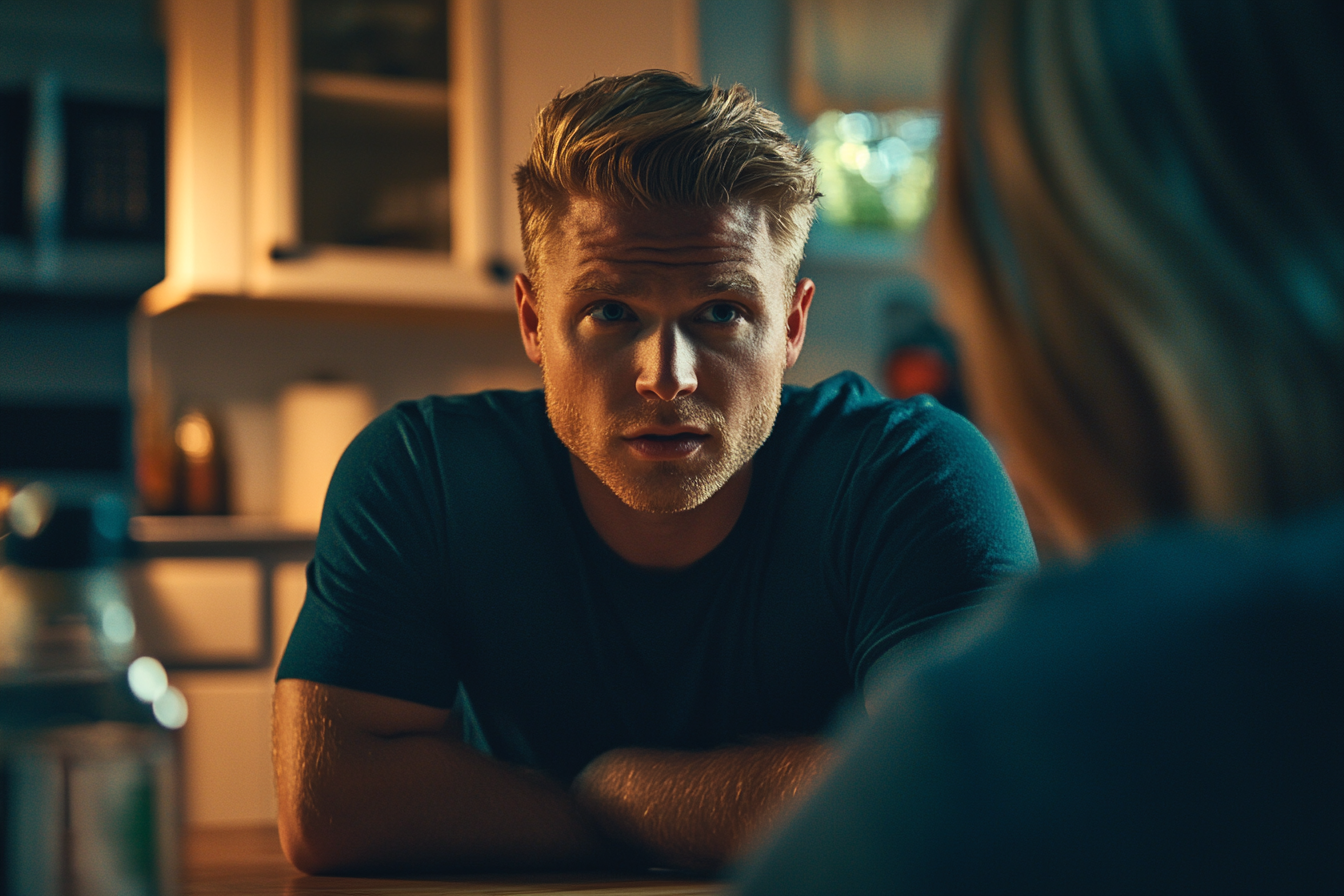 A blonde man in his 30s looking worried while talking to a woman at the kitchen table | Source: Midjourney