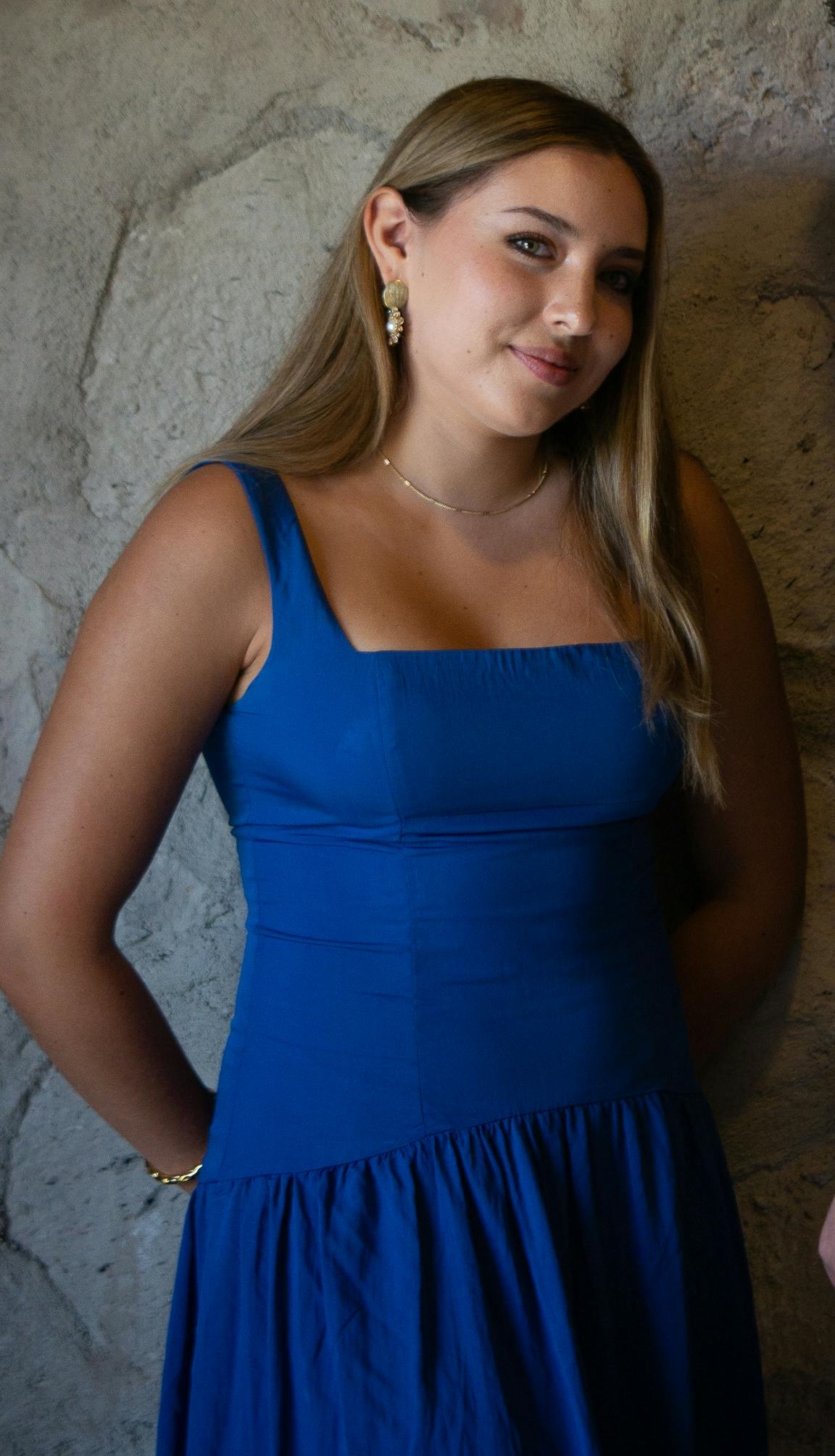 A smiling young woman in a blue dress with a necklace and earrings standing in front of a stone wall | Source: Pexels