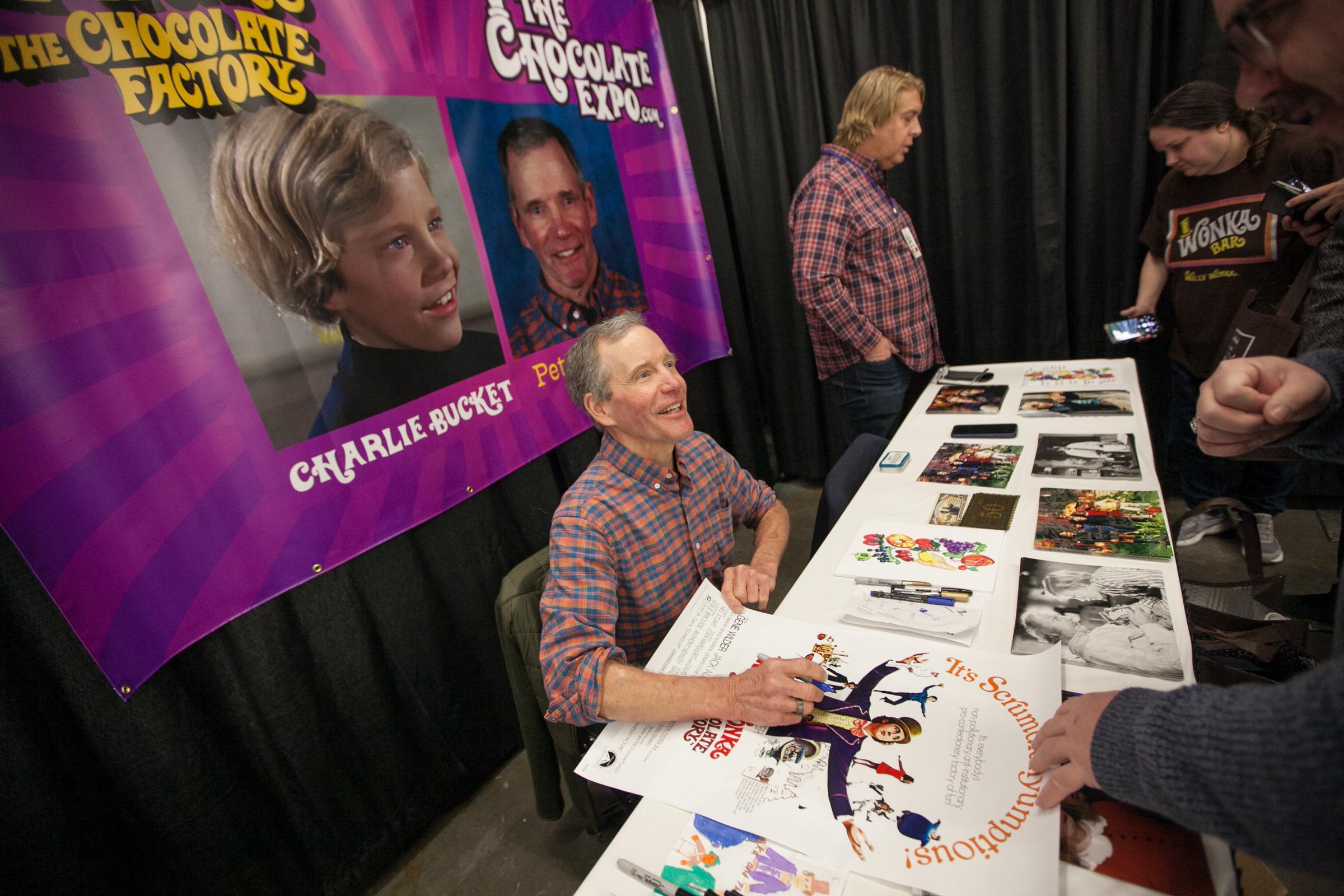 Peter Ostrum (fore left) signs an autograph for attendees during an event related to the film "Willie Wonka and the Chocolate Factory" at the Chocolate Expo in the Shriners Auditorium, in Wilmington, Massachusetts, on January 27, 2024.┃Source: Getty Images