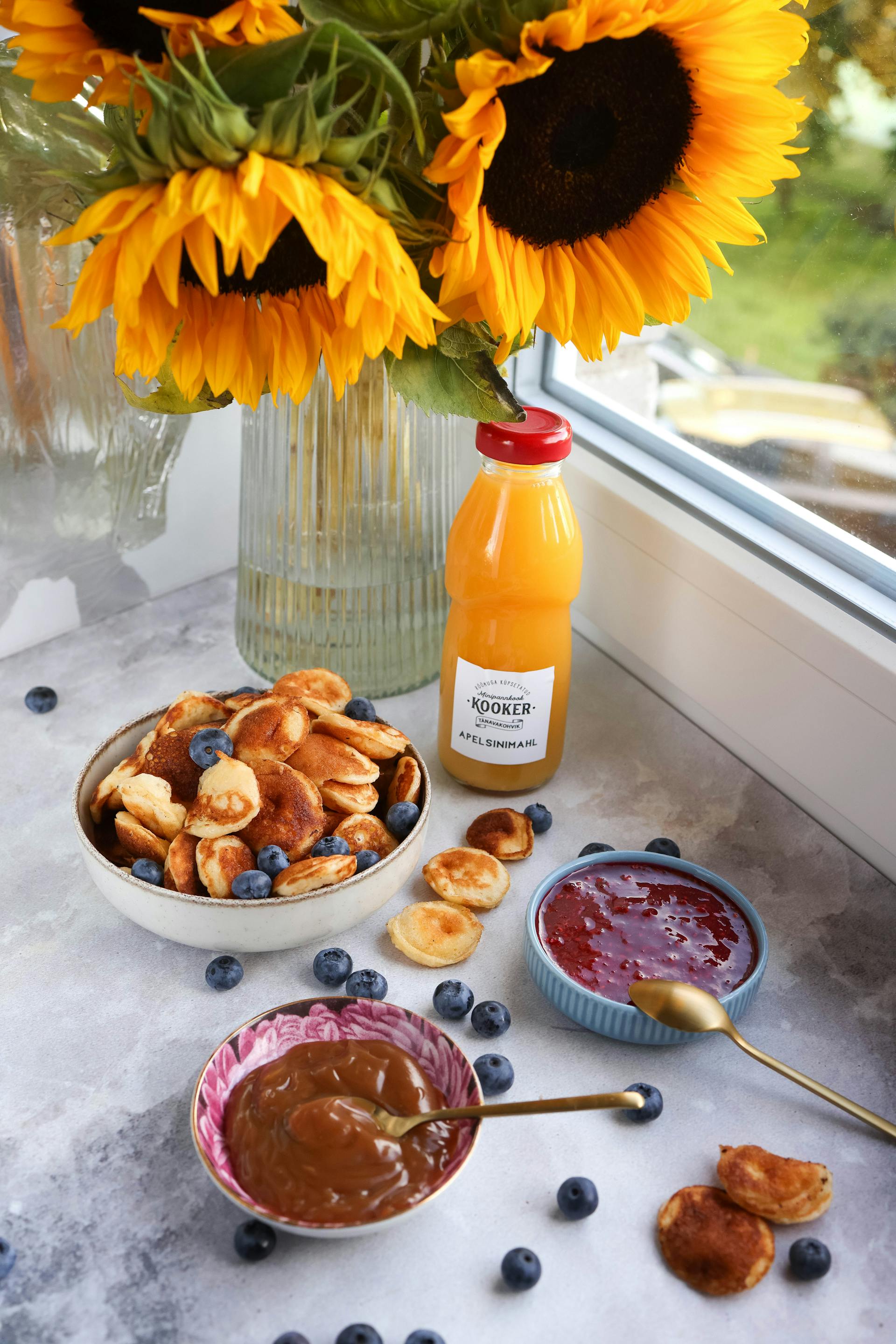 A photo showing pancakes with blueberries and fruit juice lying near a vase of sunflowers beside the kitchen window | Source: Pexels