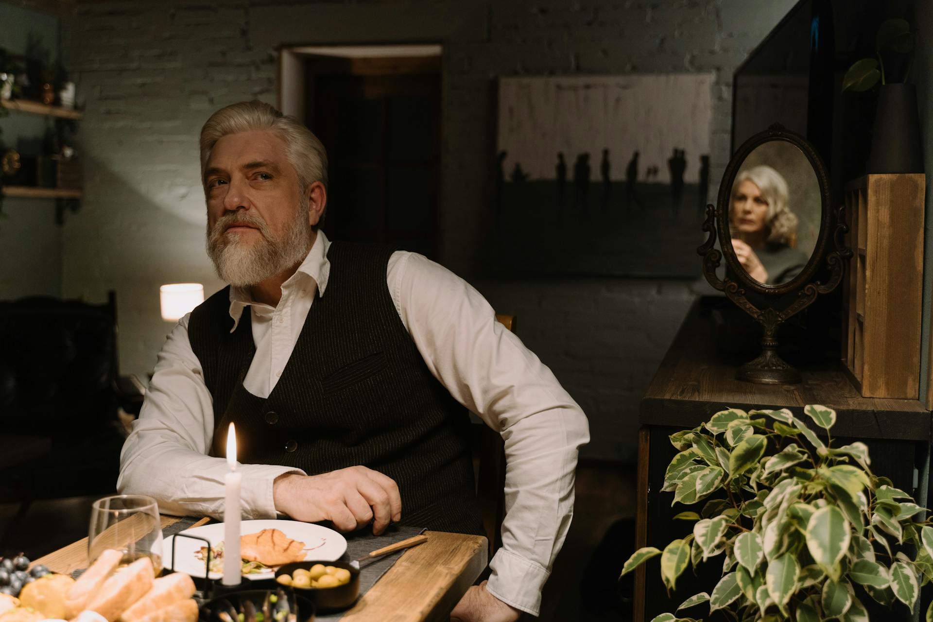 A senior man seated in a dining room while a mirror casts his partner's reflection from the table behind | Source: Pexels